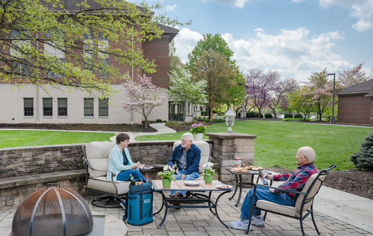 a senior couple and a man sit laughing and talking at an outdoor patio area with firepit having a picnic with beer, cheese, and snacks