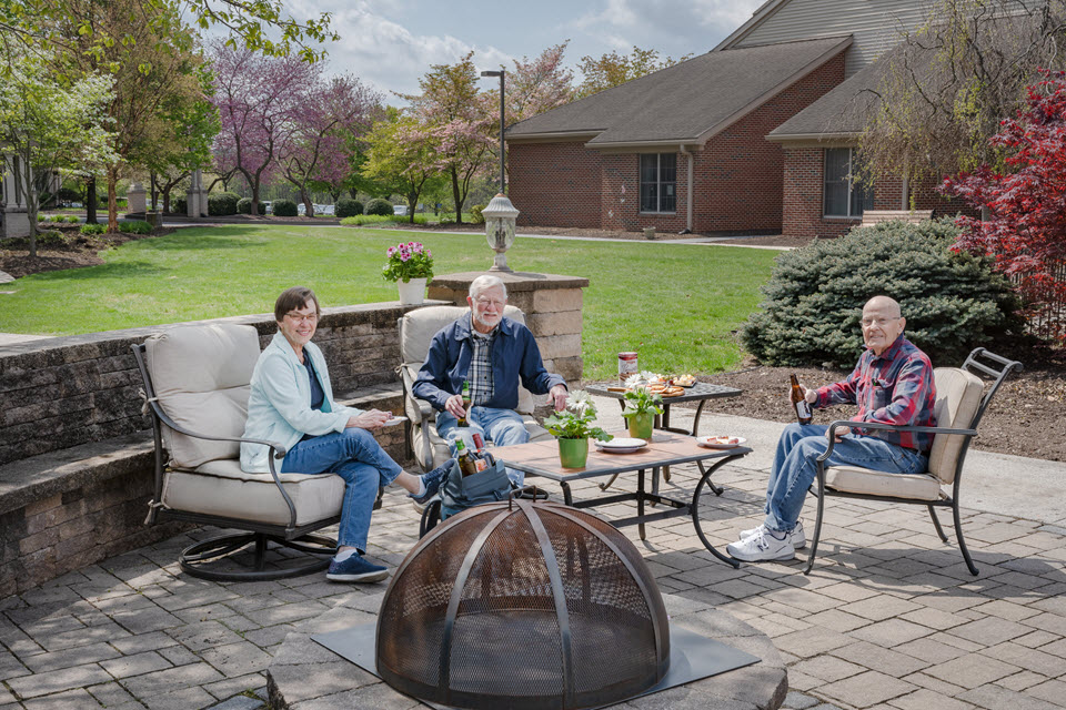 a senior couple and a man smile at camera at an outdoor patio area with firepit having a picnic with beer, cheese, and snacks