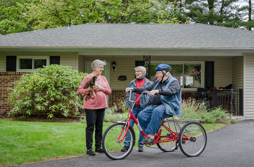 two senior women stand by a man on a three wheeled bike looking at a dog