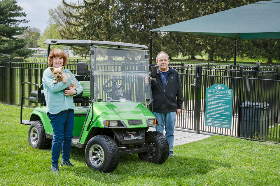 man and woman stand by green golf cart with their dog in front of bethany village dog park