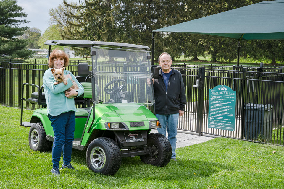 man and woman stand by green golf cart with their dog in front of bethany village dog park