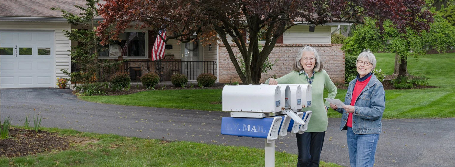 two senior women stand at mailboxes in front of cottage smiling at camera