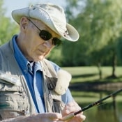 older man in hat and sunglasses by a pond, with fishing gear