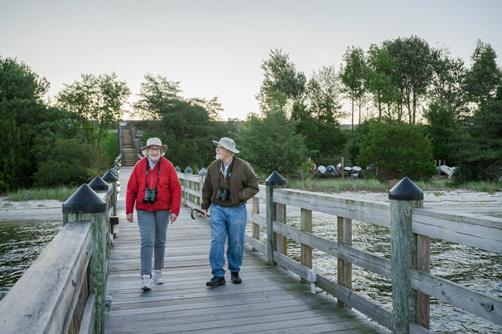 an older man and woman walking a pier with hats on and binoculars