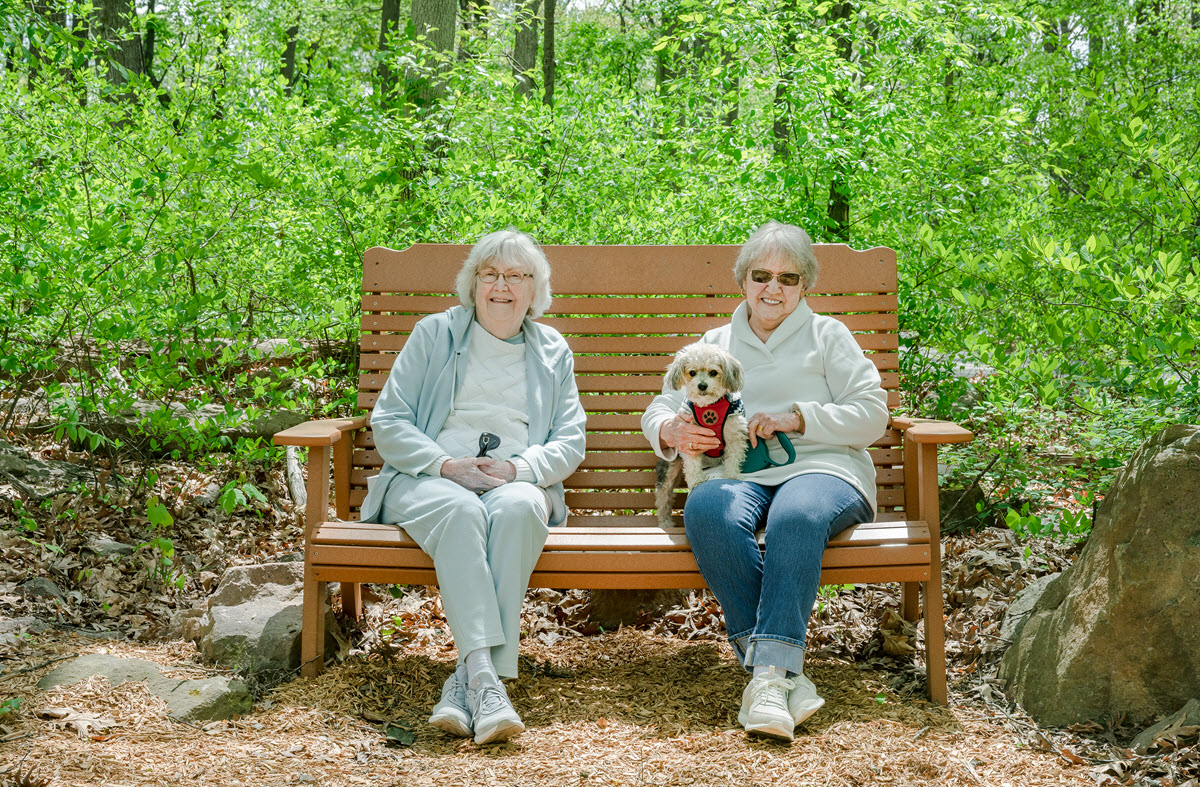 two senior women sitting on an outdoor bench, one with a small dog in her lap