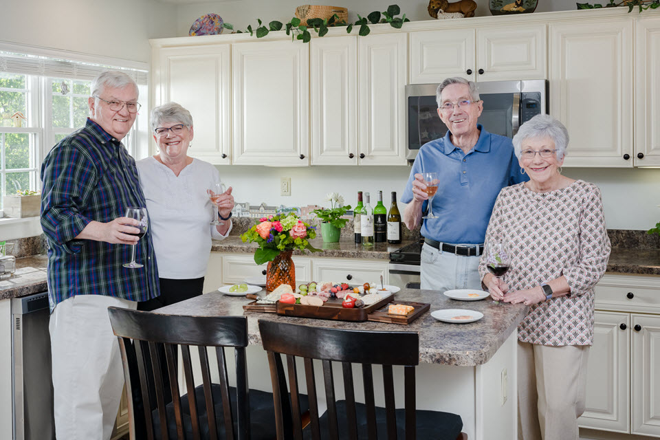two senior couples stand in a large kitchen with an island holding charcuterie and wine