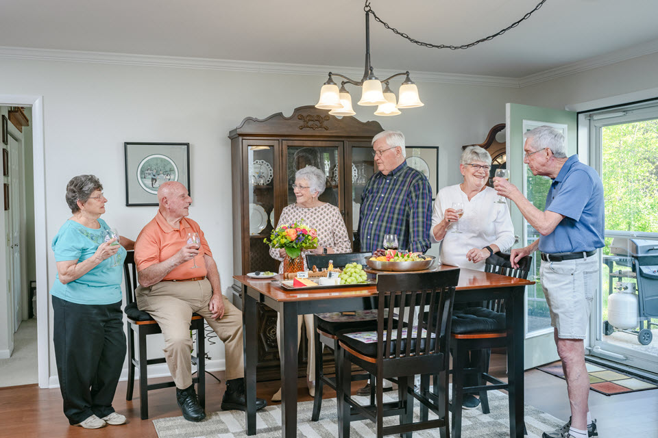 six seniors stand around a dining room table with wine and fruit, cheese platters