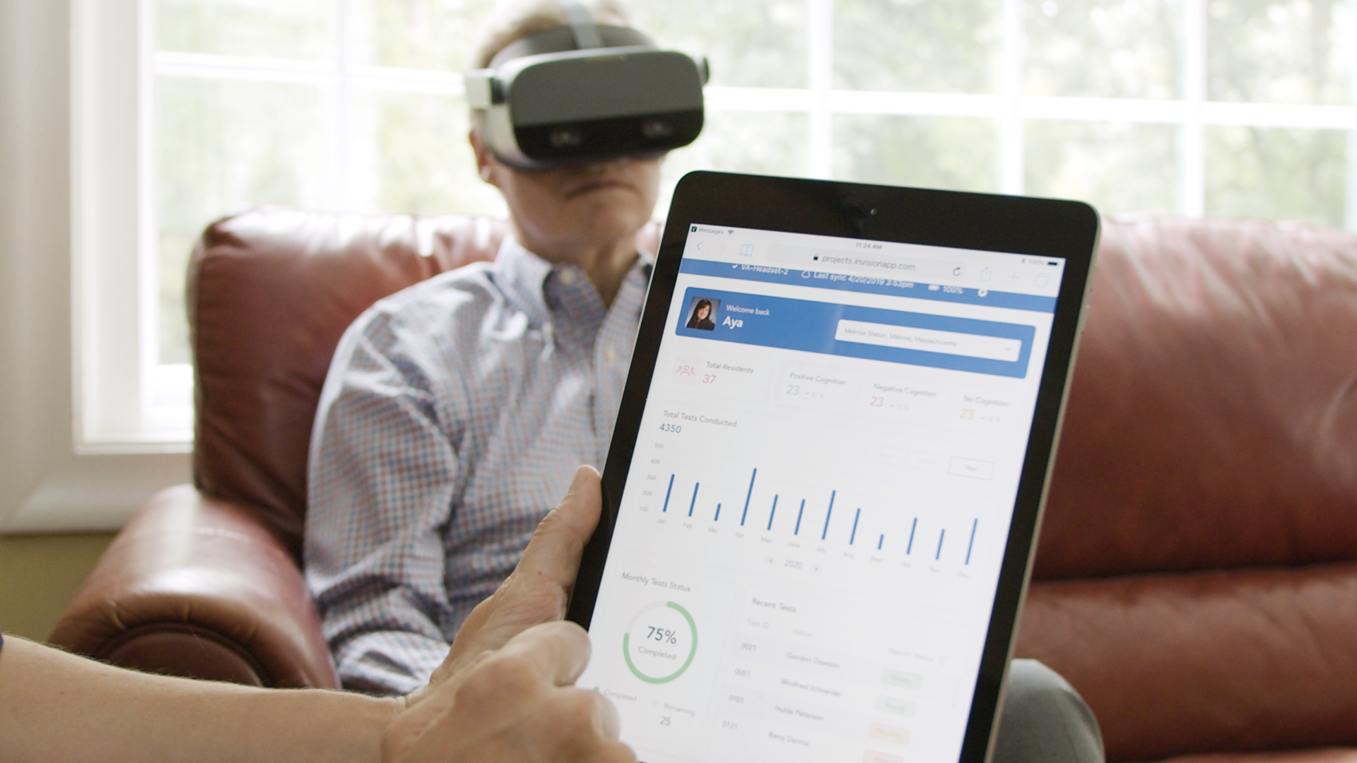 Man wearing a virtual reality headset sitting on a couch while a person holds a tablet displaying data.