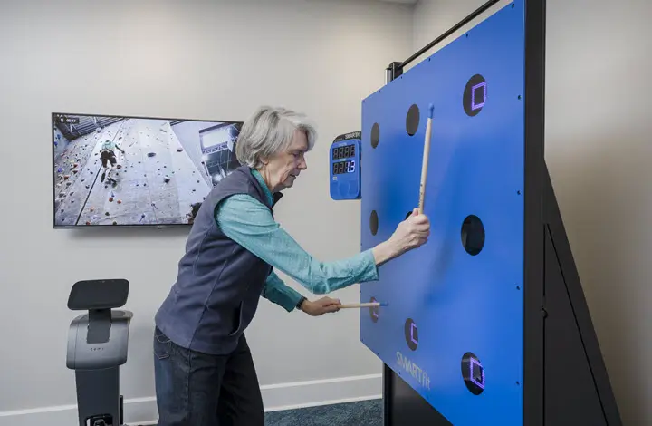 senior woman gray hair wearing a vest playing with the smart fit machine, drumstick in hand