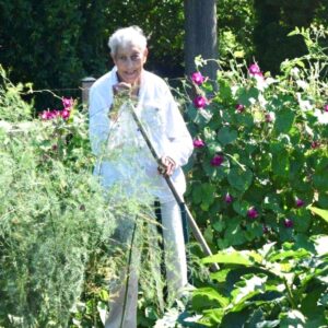 older woman standing in garden holding hoe with flowers and squash vines