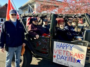 A jeep with a Happy Veterans Day sign on the side and flags on the back with four senior veterans sitting inside and one man standing beside the jeep