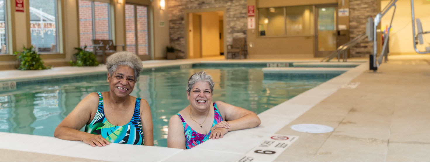 african american senior female and white female stand in corner of an indoor pool surrounded by large windows smiling at the camera