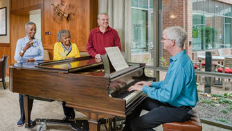 senior man sits at grand piano with two older african american women and one older white male leaning on the end having drinks and enjoying and smiling to the music