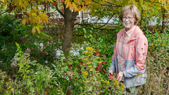senior woman in gardening gloves and pink jacket stands in wildflower garden smiling and trimming back dead plants