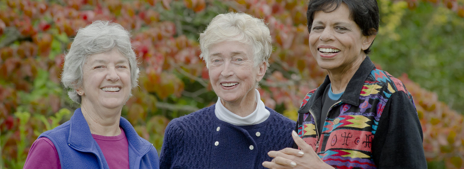 two white and one african american senior women standing outside with fall leaves in the background and smiling at the camera