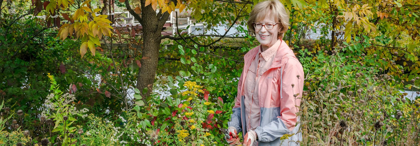 woman in pink jacket stands in wooded garden wearing gardening gloves to prune the bushes and flowers