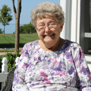 Judi Jones sits on her porch at Asbury Solomons with the Patuxent River in the backgrounds. She is wearing a floral shirt and smiling at the camera.