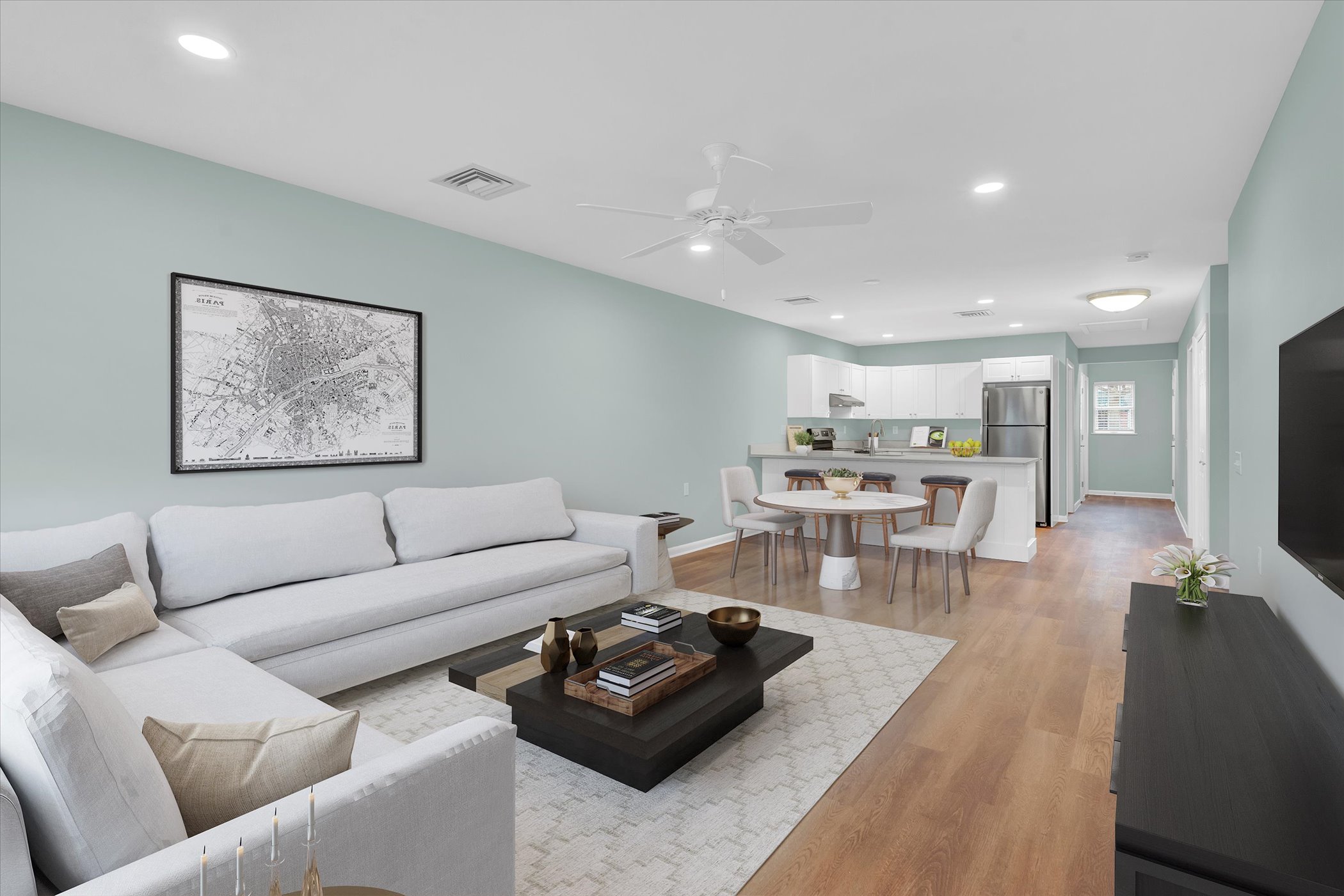 open floorplan living room with pale green walls, large window and french door, coffee table and sectional sofa with rug over hardwood floor, looking into open kitchen