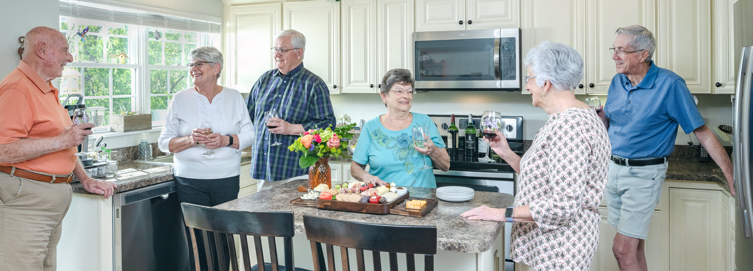 three senior couples stand in modern open kitchen having wine and cheese and laughing