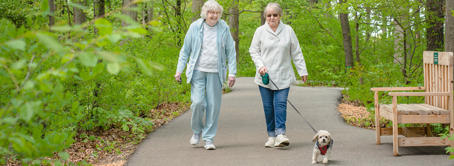 two senior women talk and laugh as they walk a dog through wooded trail