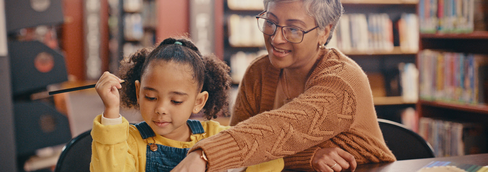 older woman sits in library with young girl helping her read