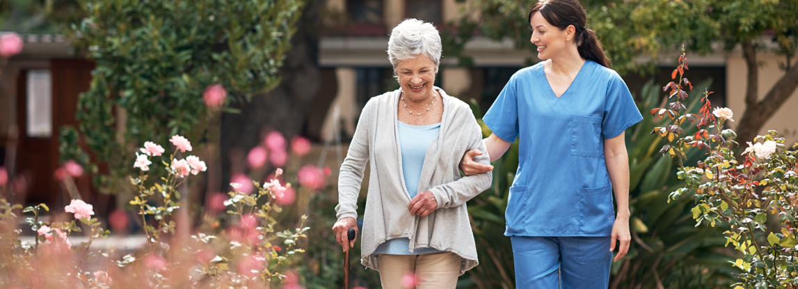 outdoor garden. elderly white woman with cane and sweater walks with white caregiver in blue scrubs
