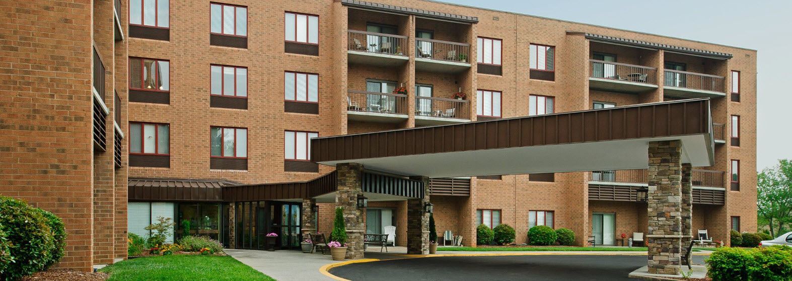 large brick apartment building with four stories and patios, green lawn, and covered entryway off of a parking lot