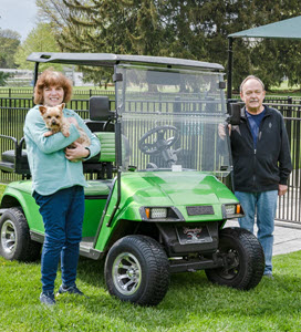 senior woman and man stand by their golf cart at Bethany Village dog park holding a brown dog