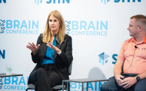woman in business suit sits in front of a backdrop that says brain conference speaking to a group