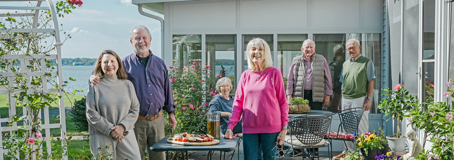 three senior couples stand on flagstone cottage patio enjoying desserts and ice tea surrounded by roses, flower pots with patuxent river in background