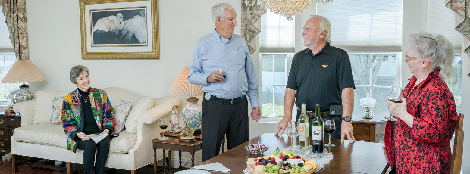 two senior couples stand in large formal living room having wine and cheese and laughing