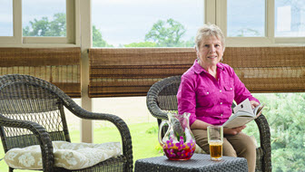 woman in pink shirt sits on her sun porch with ice team and a book with mountains in background