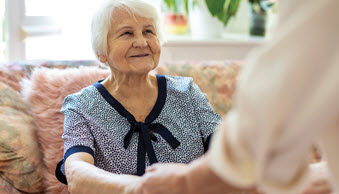 elderly woman sits on sofa smiling at a person who is reaching out her hands to help her up