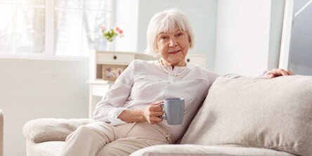 senior woman sits on white sofa smiling with a cup of coffee in her sunny apartment
