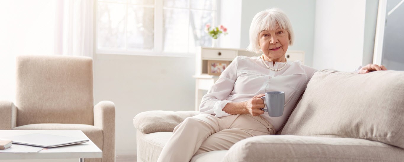 senior woman sits on sofa in bright room with cup of coffee and open window behind her