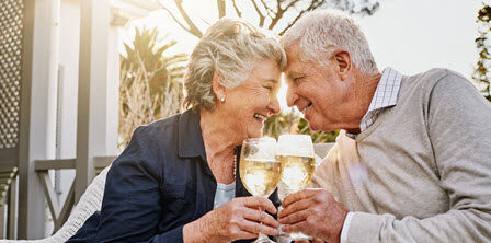 senior man and woman sit on sunporch touching foreheads and toasting with glass of wine