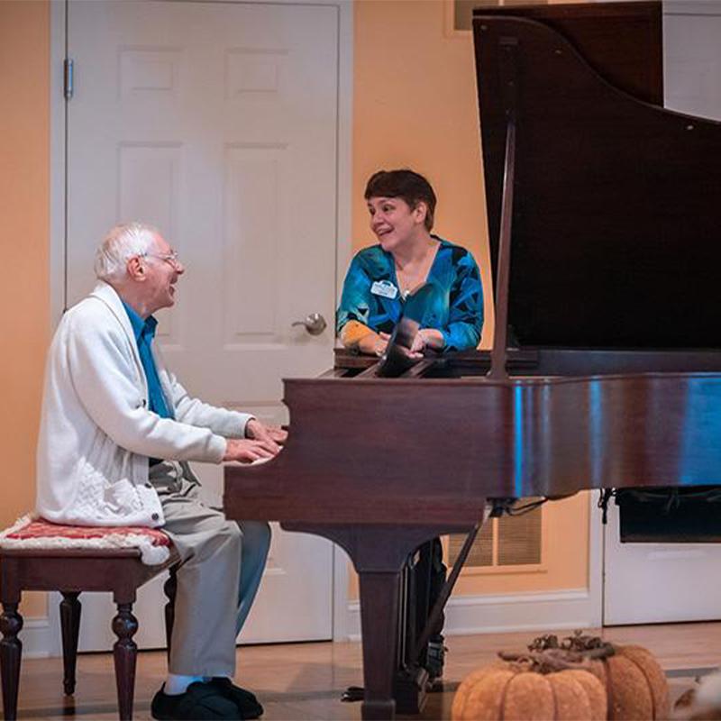senior male resident playing piano while female resident stands next to it listening