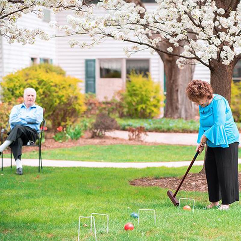 an older woman playing croquet while her husband watches her from a seated position