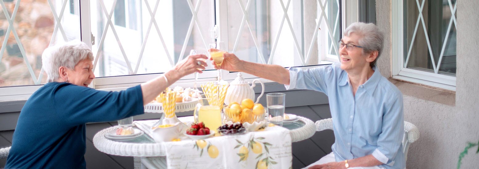 two senior women sit at wicker table in front of sunny table with fresh strawberries laughing and toasting with a mimosa