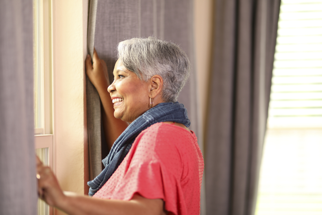 African descent, senior adult woman looks out her window on a sunny summer day.