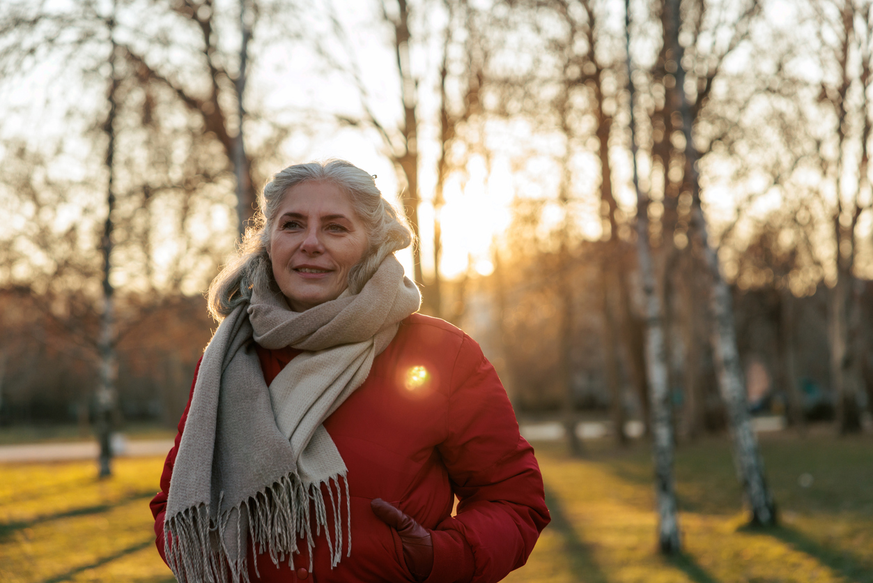 Mature woman wearing a red jacket and warm scarf walking through a park at sunset. Golden sunlight shines through the bare trees, creating a soft backlit glow and long shadows across the grass.